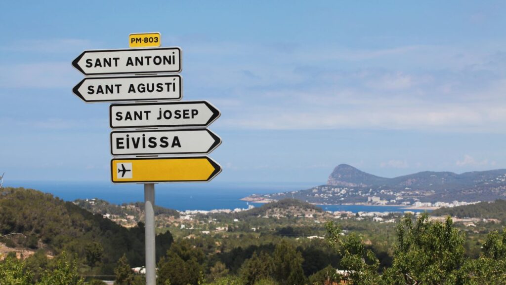 Wegweiser auf Ibiza mit den Zielen Sant Antoni, Sant Agustí und Eivissa, im Hintergrund die Küste – inspiriert von Ibiza Restaurant Geheimtipp.